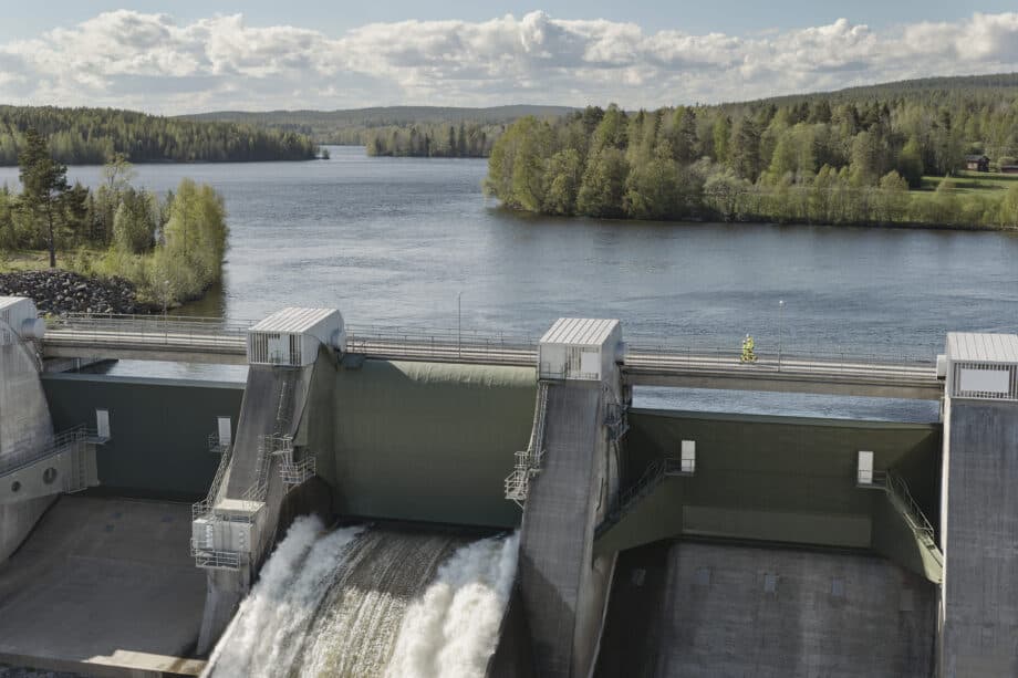 En damm som släpper ut vatten i en flod, med ett skogslandskap och en molnig himmel i bakgrunden.